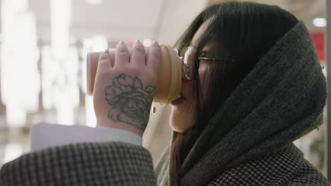side view of lady with tattooed hand wearing thick scarf and glasses sipping coffee from paper cup in softly lit modern indoor space with white lights glowing in background