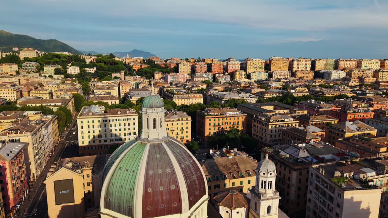 Drone orbits above the dome of Genoa’s Cathedral of San Lorenzo during the golden hour, showcasing the city’s colorful buildings, hillside housing, mountains, and sea in the distance