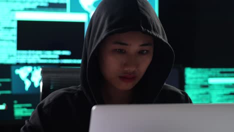 close up of asian female hacker in a hood showing hand palm to camera while works on a laptop with maps and data on display screens of multiple monitors in a dark office room