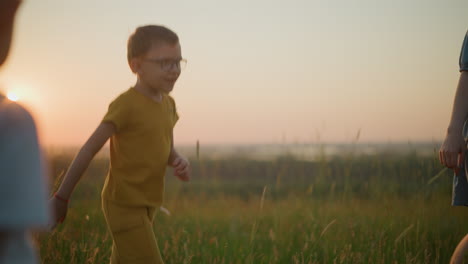 primer plano de un niño con ropa y gafas amarillas, bailando felizmente en un campo cubierto de hierba durante la puesta de sol. una mujer con un vestido azul, sosteniendo un teléfono, es parcialmente visible en el fondo