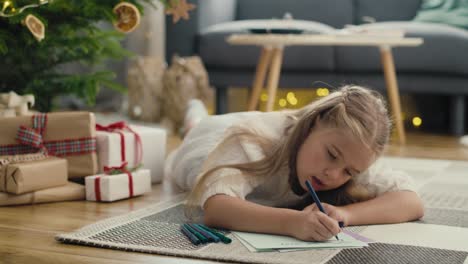 little caucasian girl lying on carpet next to christmas tree and writing a letter to santa claus.