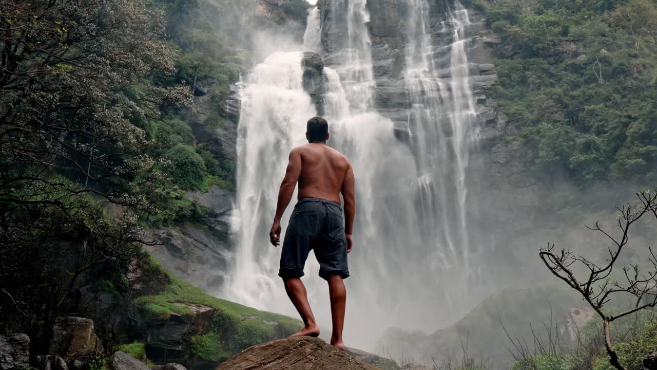 A man stands near the base of Bomburu Ella Waterfalls, surrounded by mist, rocks, and dense greenery in the highlands of Nuwara Eliya, Sri Lanka.