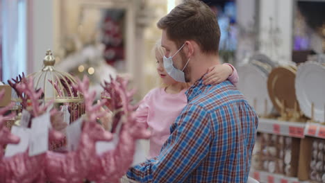 a father in a medical mask with his daughter in a shopping center choose home decorations for christmas. on christmas eve preparing to decorate your home during the coronavirus pandemic