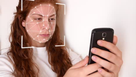 Close-up-of-a-young-woman-with-red-hair,-with-a-white-shirt-on-a-white-background,-while-holding-a-phone-in-front-of-her-face-with-facial-recognition-high-tech-animation-with-a-glow-effect-on-her-face