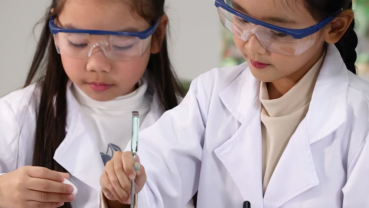 Kaksi keskittynyttä aasialaista koulutyttöä, suojalasit ja laboratoriotakit päällä, käyttävät pipettiä kemiassa.