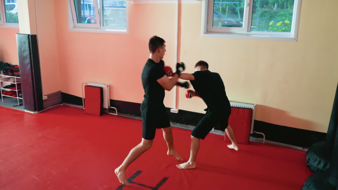 Wrestlers facing off barefoot in gym training session, wearing gloves and black outfits, grappling combat with guarded stances on red mat, showing discipline, intensity, agility, power