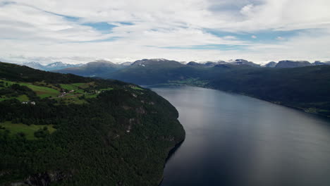 aerial shot, panning over a wooded hillside, revealing a fjord stretching out into the distance, winding between the steep hills and snow capped mountains of ålfotbreen park in norway