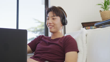 happy asian male teenager wearing headphones and using laptop in living room