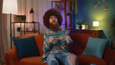 a man in a 70s retro outfit celebrates while sitting on a couch in his living room