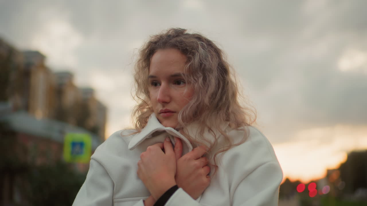 Lady feeling cold covering herself with white coat while standing outdoors under cloudy sky, soft blurred background featuring urban buildings, trees, and soft glow of evening light in the city