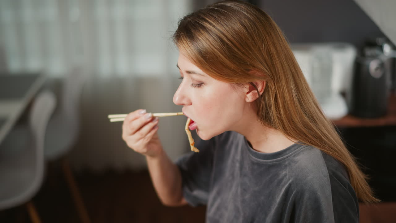 Hungry girl enjoys breakfast using chopsticks, sitting in bright modern kitchen with soft natural light and blurred dining chair in background