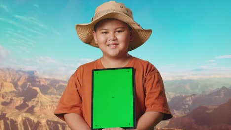 asian boy with a hat and binoculars smiling and showing green screen tablet. boy researcher examines something while traveling at the top of mountain, travel tourism adventure concept, close up