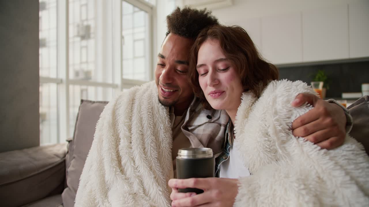 A happy young man with Black skin color in a checkered cream shirt sits on a modern sofa wrapped in a white woolen blanket with his young girlfriend who drinks tea from a black glass they hug and spend time together in a modern apartment