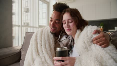 A-happy-young-man-with-Black-skin-color-in-a-checkered-cream-shirt-sits-on-a-modern-sofa-wrapped-in-a-white-woolen-blanket-with-his-young-girlfriend-who-drinks-tea-from-a-black-glass-they-hug-and-spend-time-together-in-a-modern-apartment