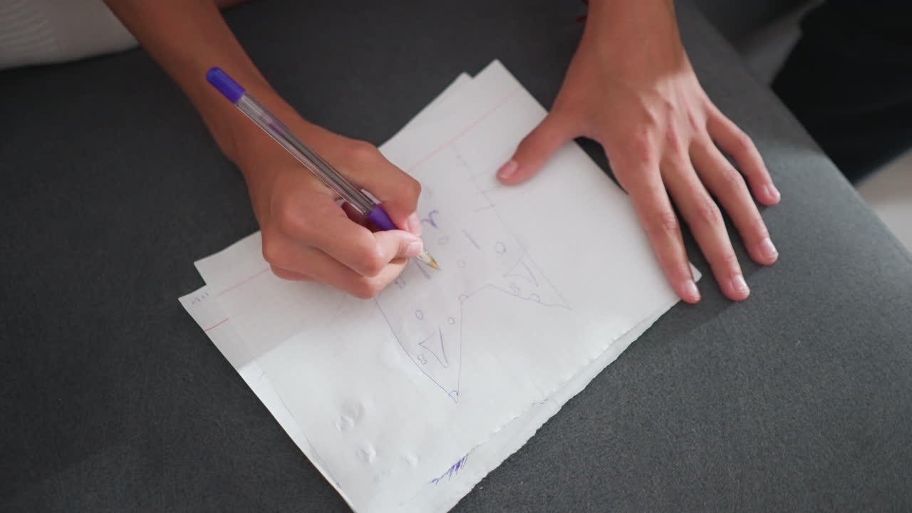 Vista desde arriba de una persona sentada en un sofá dibujando formas creativas con un bolígrafo sobre papel, mostrando el movimiento y el detalle de la mano mientras trabaja en el dibujo en un ambiente hogareño relajado