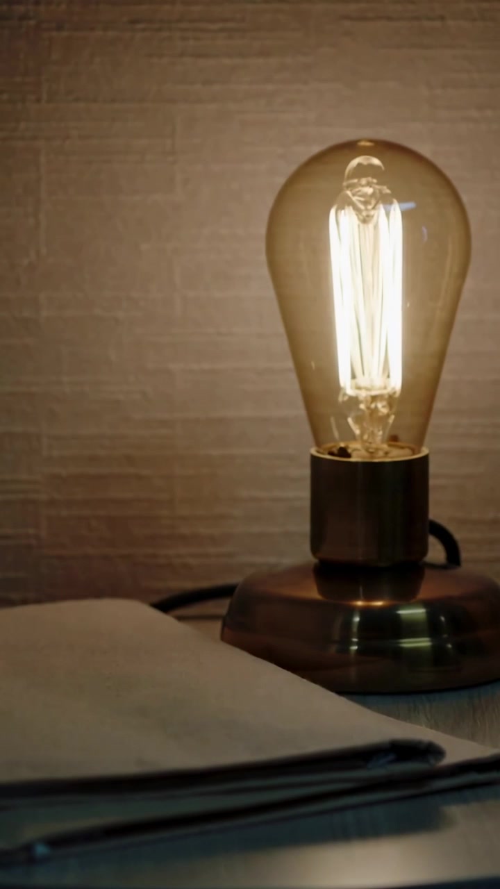 Video verticale: La telecamera zooma avvicinando una lampada Edison sulla scrivania, proiettando un caldo bagliore su un quaderno.
