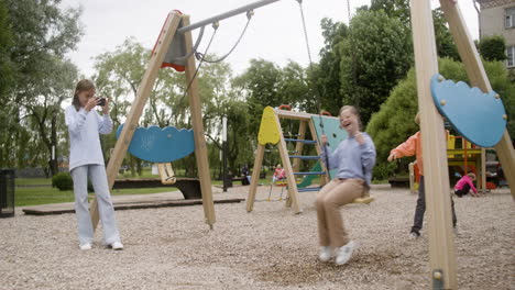 little girl with down syndrome wearing hoodie swinging on a swing in the park on a windy day. her male friend pushing her while her female friend take a photo