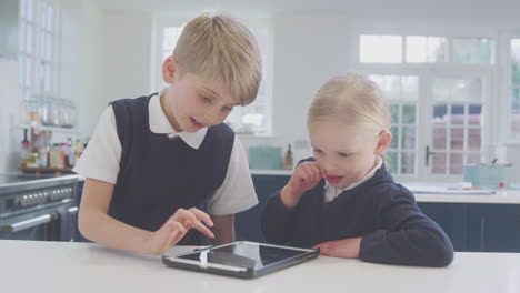 two children wearing school uniform in kitchen playing with digital tablet on counter