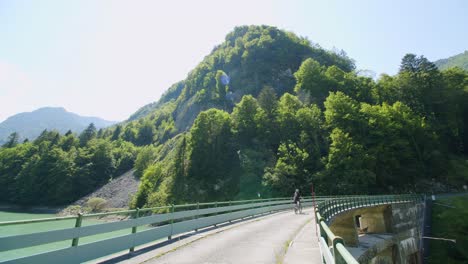 mountain biker rides across a dam with a tall mountain in front of him