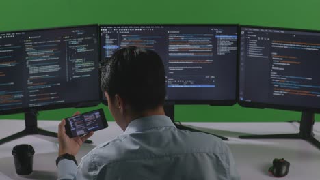 back view of asian man developer working with mobile phone while write code with multiple computer screens in green screen studio