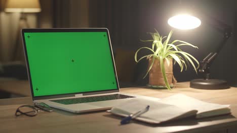 laptop computer showing green chroma key screen stands on a desk in the living room. in the background cozy living room in the evening with warm lights on