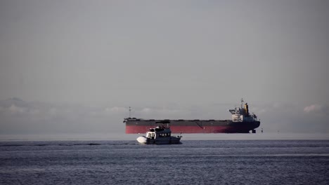 Giant-empty-Bulk-carrier-with-dropped-anchor-waiting-for-orders-while-a-small-white-yacht-is-passing-on-the-foreground-on-a-cloudy-day