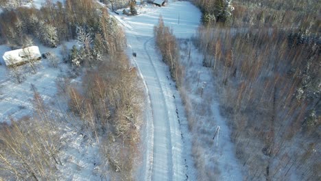 Aerial view of a serene winter countryside featuring snow-covered trees, wooden houses, and a winding road through a frosty forest.