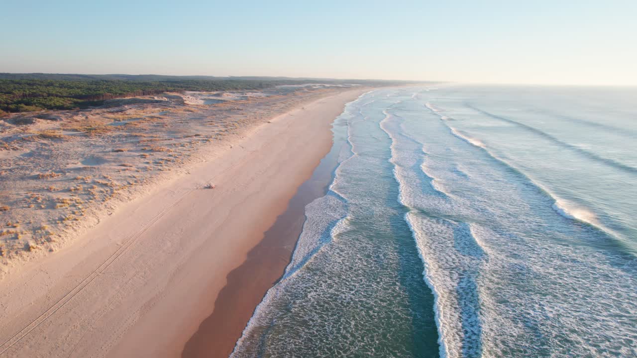 Drone følger kystlinjen langs en sandstrand med rullende havbølger