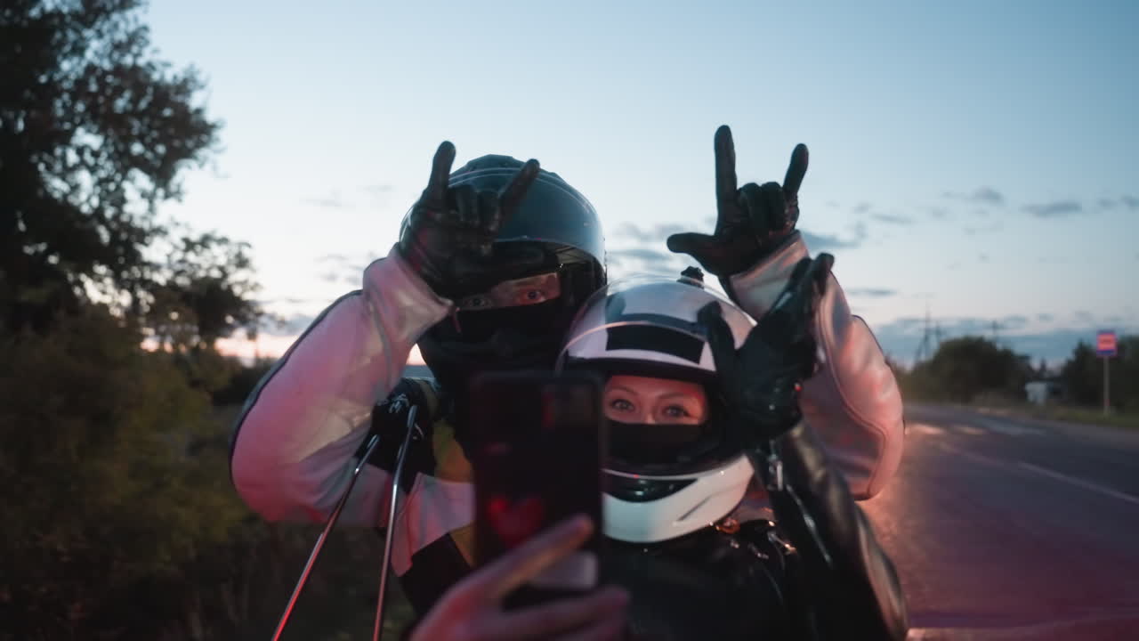 Motorcycle riders wearing helmets and leather jackets stand on roadside during sunset as woman holds smartphone taking photo with man beside her while car passes in background