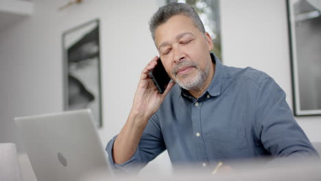 Älterer-Mann-Mit-Gemischter-Abstammung,-Der-Am-Tisch-Mit-Dem-Smartphone-Spricht-Und-Einen-Laptop-Benutzt,-Von-Zu-Hause-Aus-Arbeitet,-Zeitlupe