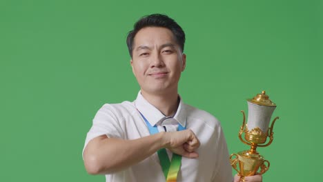 close up of asian man with a gold medal and trophy touching his chest then smiling and pointing to camera being proud of himself on green screen background in the studio