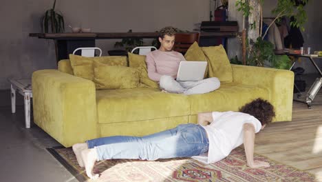 Curly guy relaxing on sofa with laptop while his boyfriend exercising on the floor