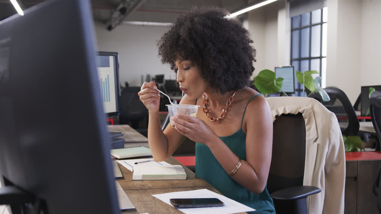 feliz mujer de negocios casual biracial sentada en el escritorio, comiendo y mirando la computadora portátil, cámara lenta