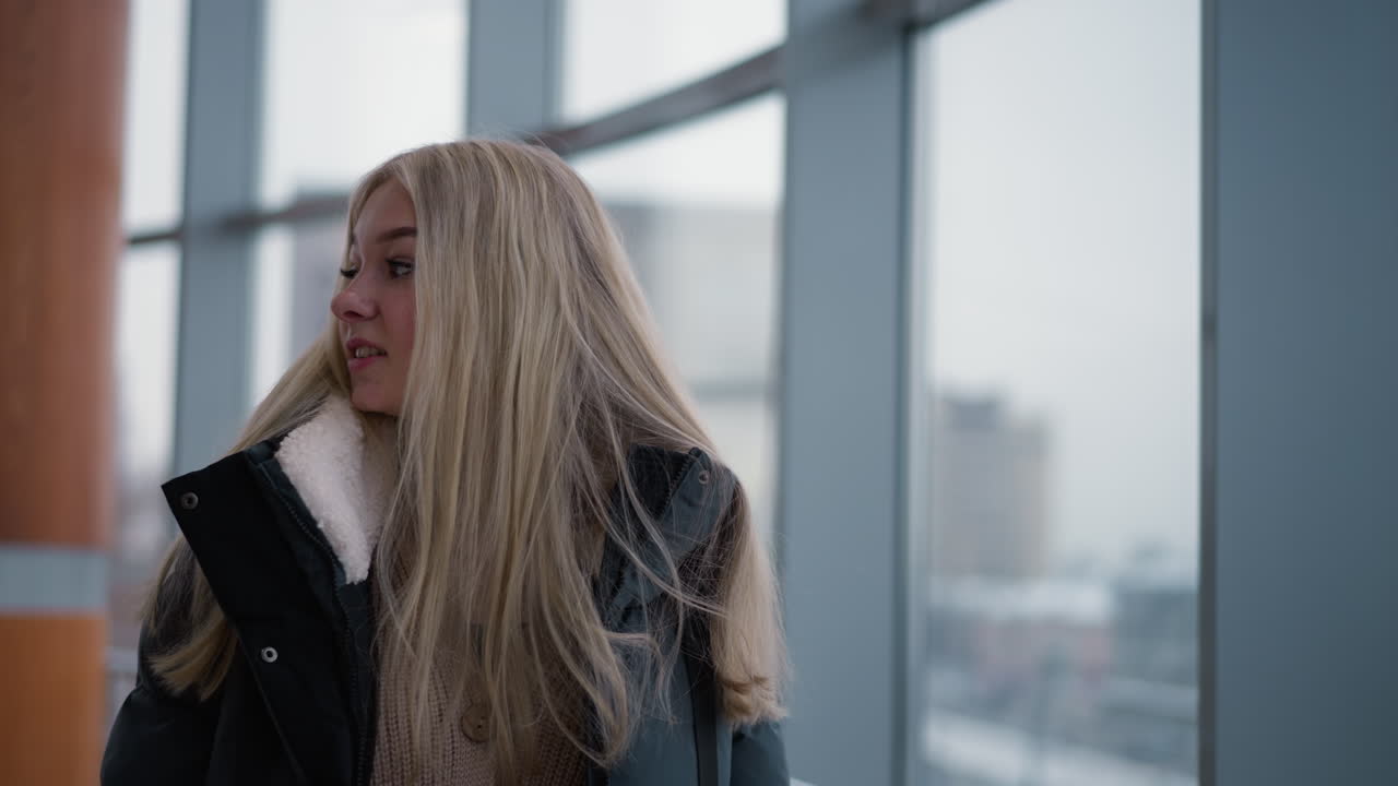 검은 겨울 재을 입은 젊은 여성은 유리 창문을 통해 도시 풍경과 건물의 흐릿한 시야를 바라보고 있으며, 도시 환경에서 편안하고 상하는 느낌을 받습니다.