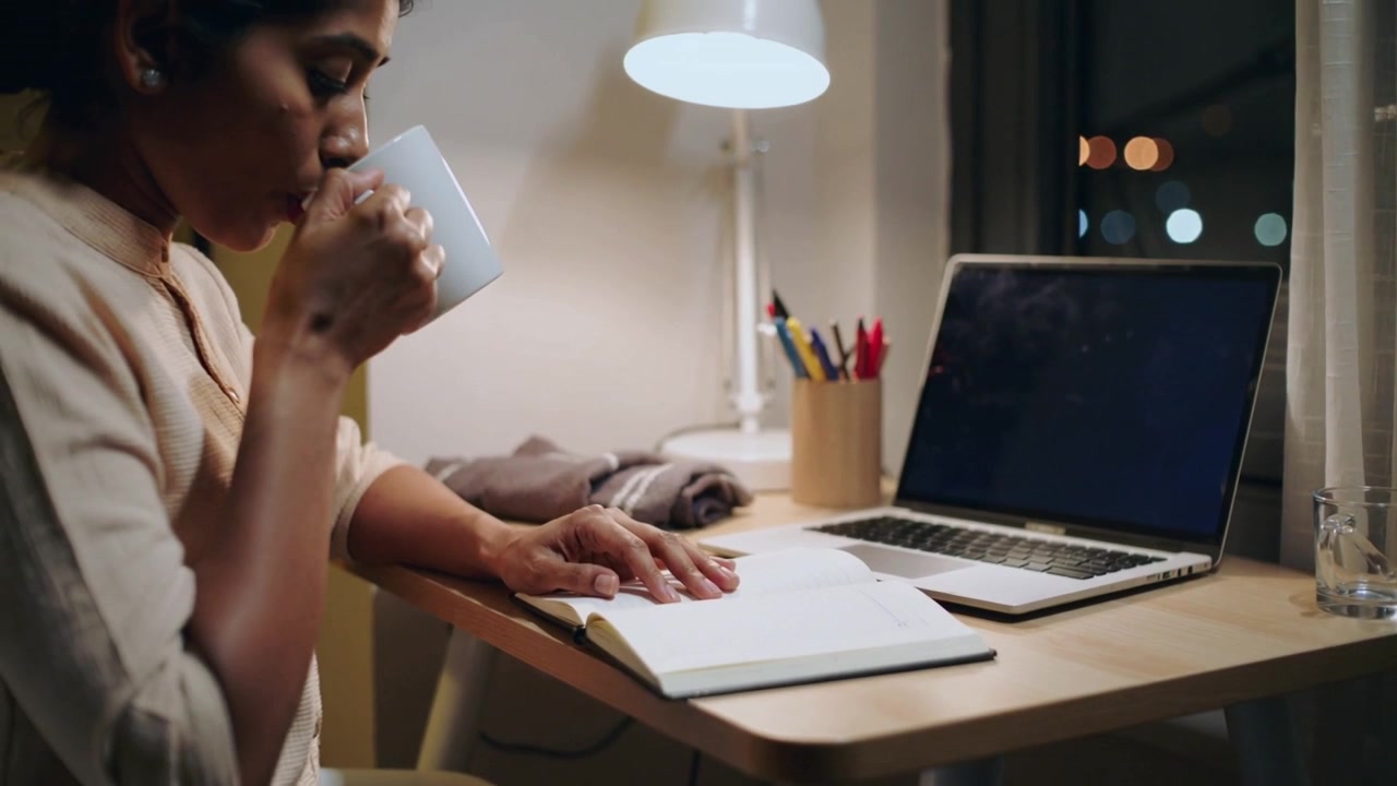 Reading notebook, woman in light sweater sipping mug at desk by window, lamp glowing, copy space