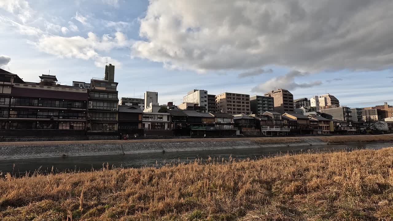 Traditional Wooden Houses Along the Kamo River in Kyoto Framed by Golden Grass and Dramatic Afternoon Clouds