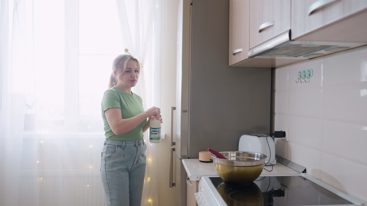 Young lady in casual outfit opens fridge door to get milk in modern bright kitchen, sunlight streaming through curtain while stainless bowl with whisk rests on stove countertop