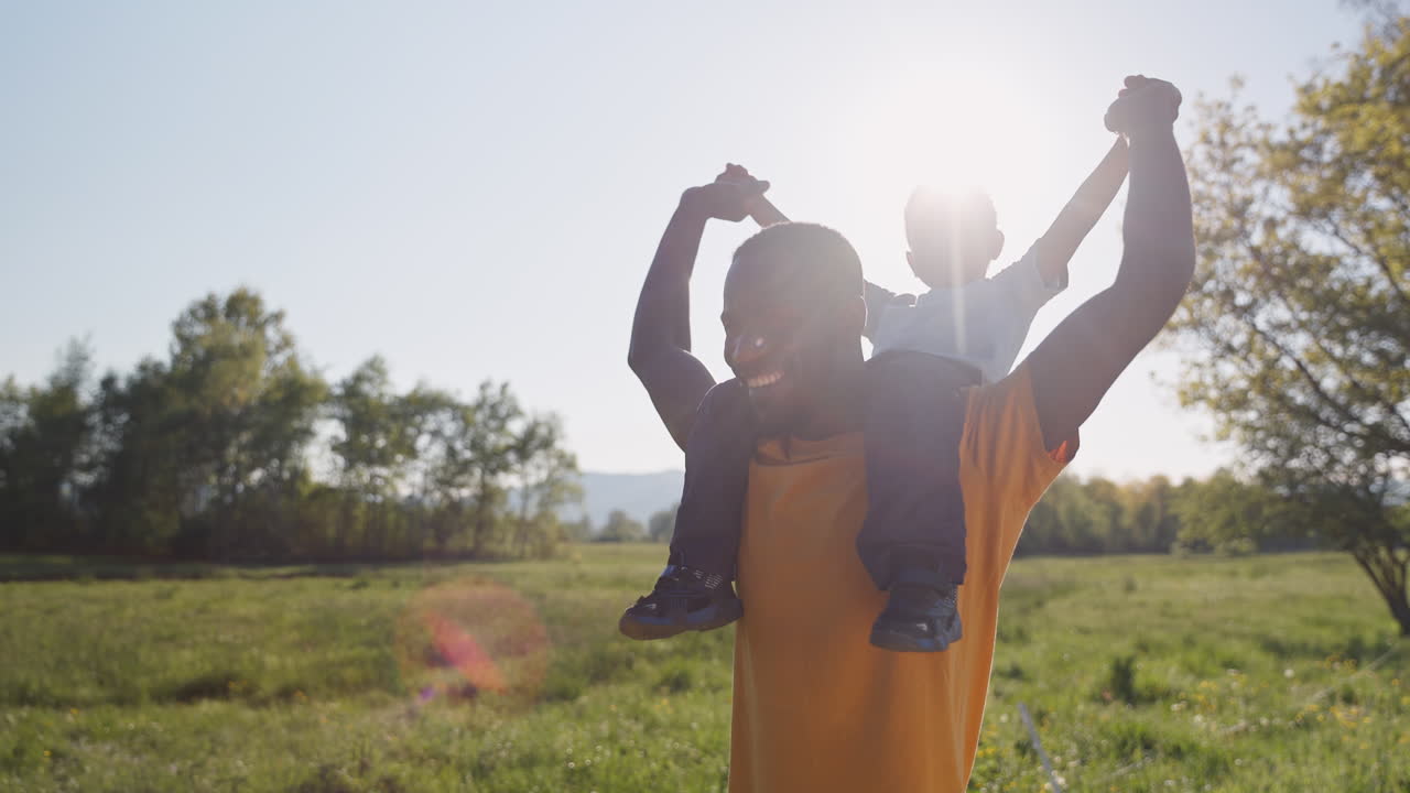 Cheerful father walking through a grassy field with his laughing son on his shoulders both enjoying