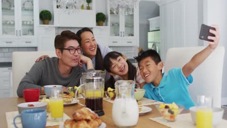 happy asian parents in kitchen having breakfast with son and daughter, son taking group selfie