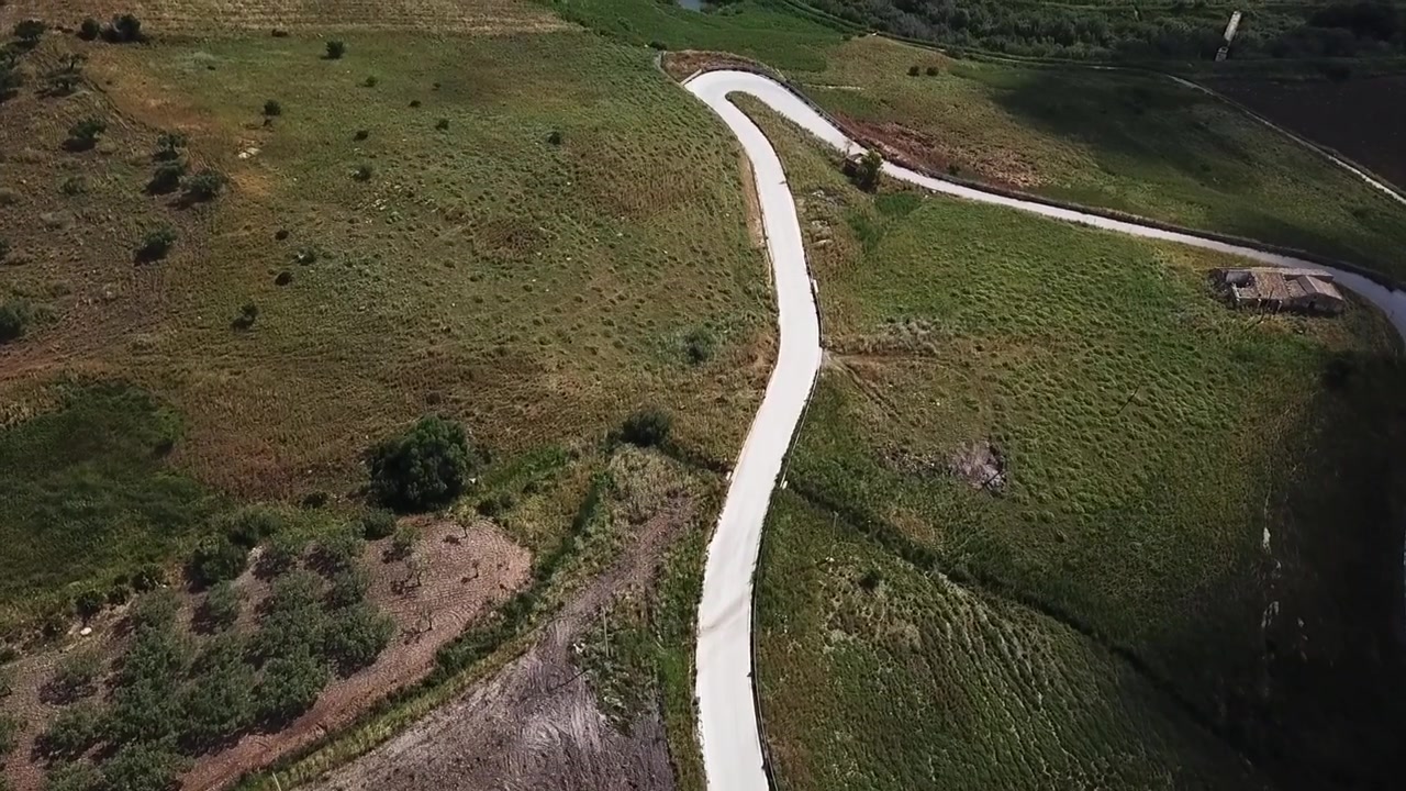 Aerial drone view of a winding, curving road cutting through dry, grassy hills and farmland in the rural Sicilian countryside, Italy, with scattered trees and a rustic barn in the distance.