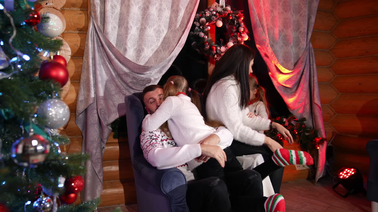 Lovely family at home at Christmas. Pretty little girls embracing their parents. Cheerful parents and cute children at the x-mas tree in the living room.