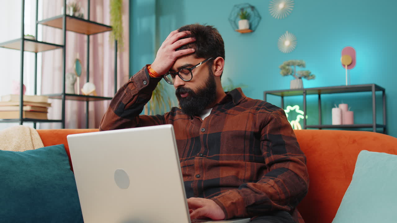 hombre indio molesto sentado en el sofá de casa usando portátil teniendo ansiedad estrés pérdida mal fallo virus de noticias