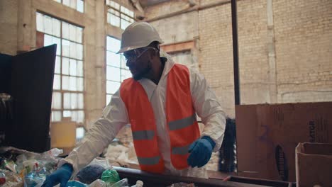 Un-Hombre-De-Piel-Negra-Con-Uniforme-Blanco-Y-Chaleco-Naranja-Y-Guantes-Protectores-Azules-Clasifica-La-Basura-Y-Las-Botellas-De-Plástico-Según-El-Color-En-La-Gran-Planta-De-Reciclaje-De-Residuos.