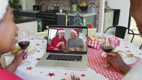African-american-couple-with-wine-using-laptop-for-christmas-video-call-with-happy-family-on-screen