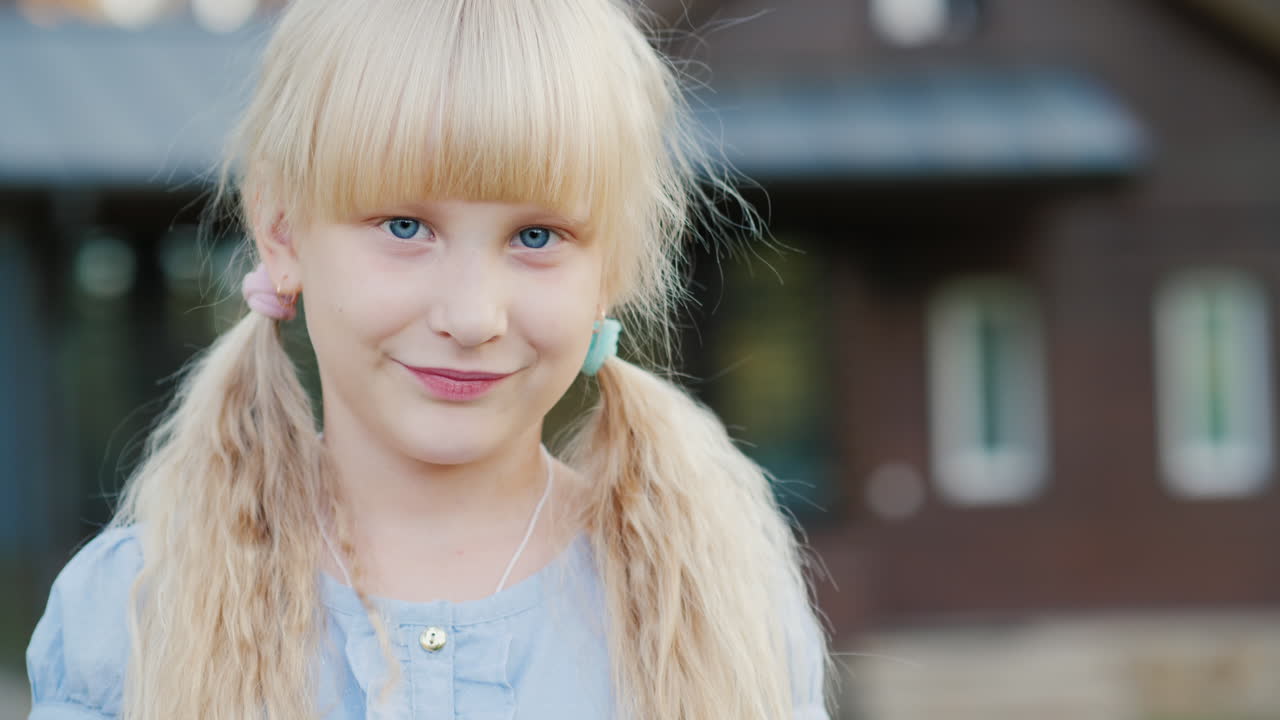 портрет белокурой девочки 6 лет, улыбающейся в камеру, она стоит на фоне своего дома