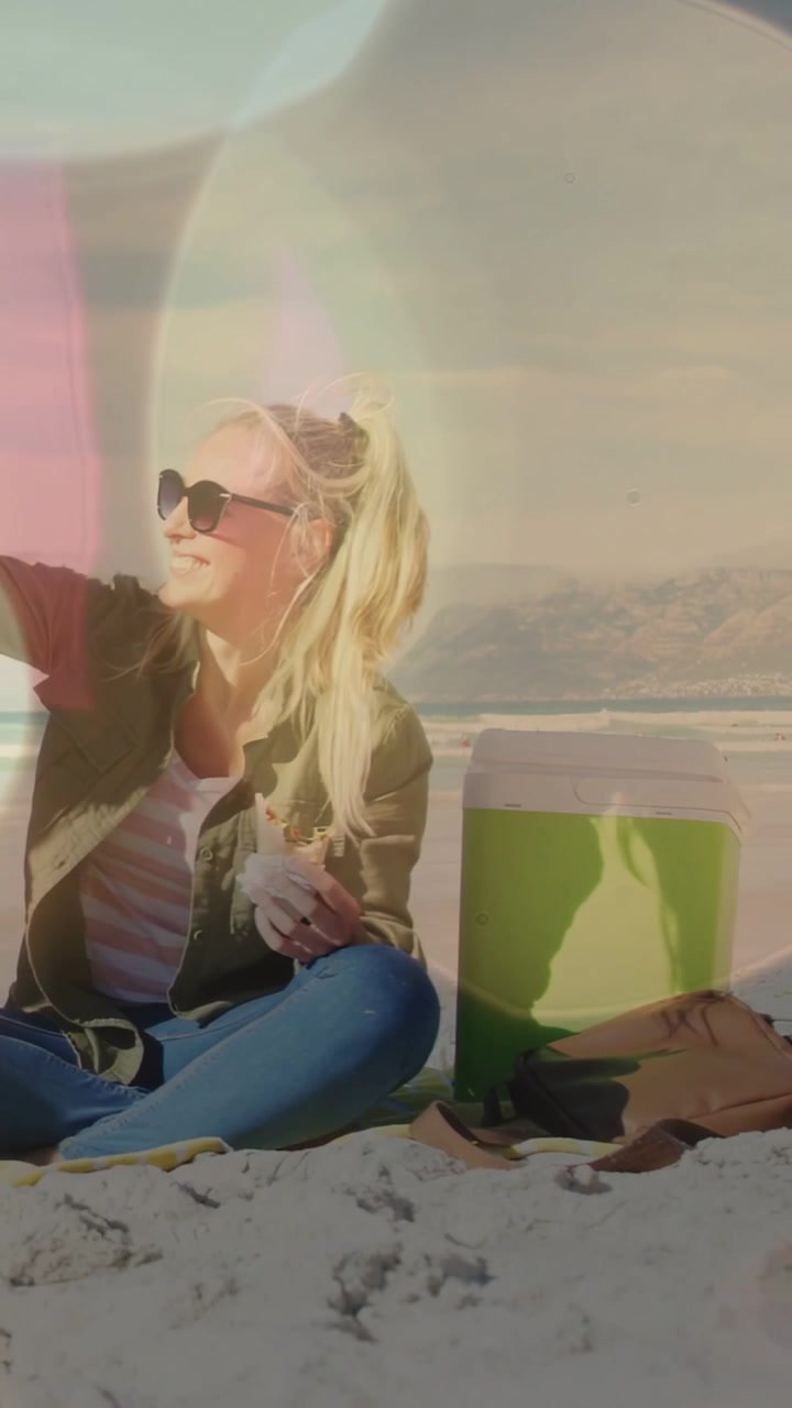 Video verticale: Donna in spiaggia che reagisce a un segnale, condivide uno spuntino, sorseggia da una tazza rossa, con bagliori dell'obiettivo.