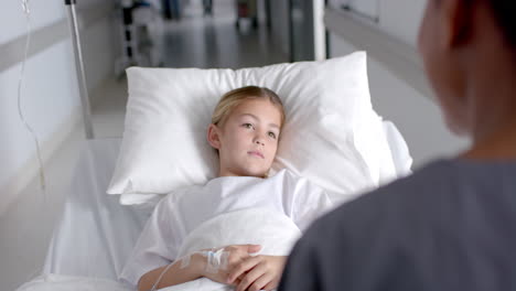 a young caucasian girl lies in a hospital bed, talking to a nurse