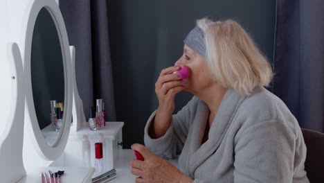 Old-senior-woman-grandmother-taking-care-of-skin,-applying-anti-wrinkle-foundation-with-sponge