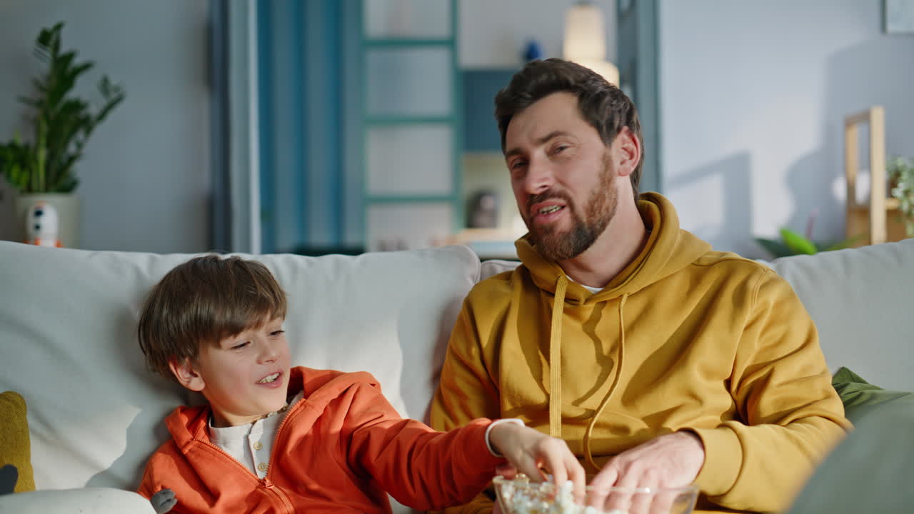 Far og søn spiser popcorn og nyder en ubekymret familiefjernsynsaften, siddende på sofaen i nærbillede.