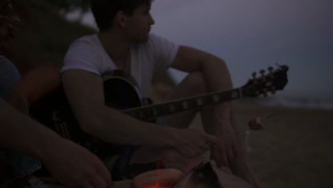 close up view of sausages grilled on the beach fire. group of young and cheerful people sitting by the fire on the beach in the